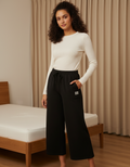 Woman wearing a white long-sleeve top and black Plazzo in a bedroom setting.