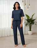 Woman wearing navy blue pajamas standing in a room with white curtains and a plant.